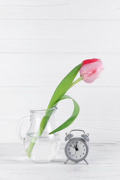 Free photo gray alarm clock near the transparent glass jug with pink tulip against white wooden desk