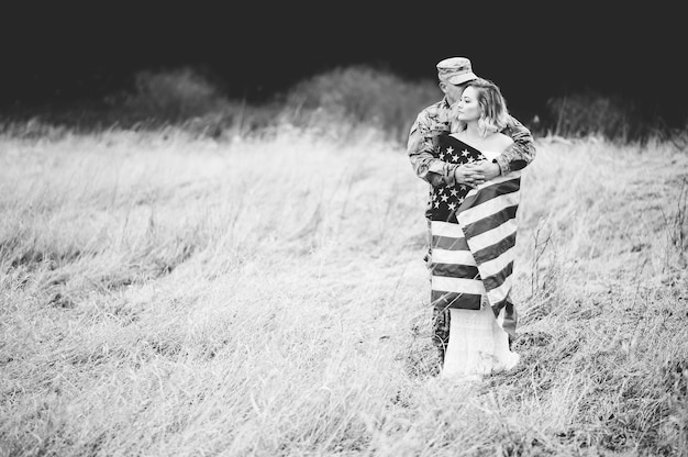 Grayscale shot of an American soldier hugging his wife while she is wrapped with an American flag