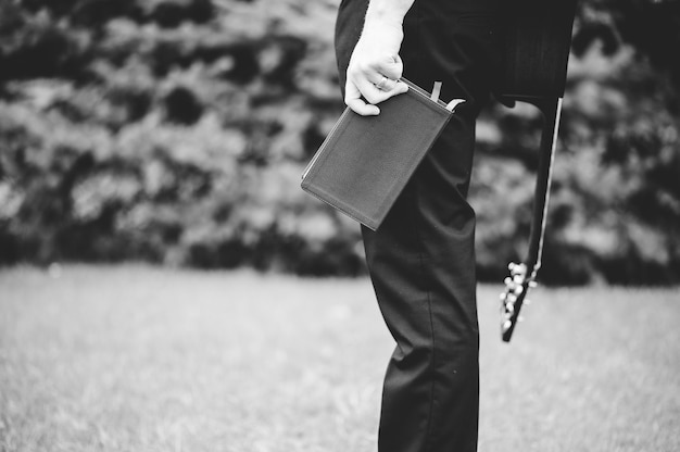 A grayscale shot of a male holding the bible and a guitar on his back