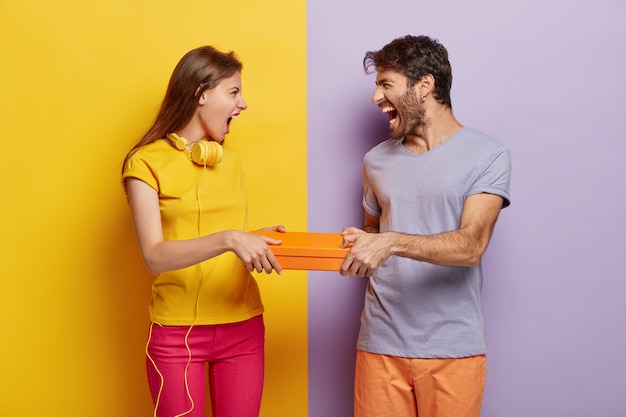 Free Photo greedy female and male cannot share box, both hold orange package, shout at each other, have annoyed expressions, wear vivid colorful clothes, stand against two colored background.