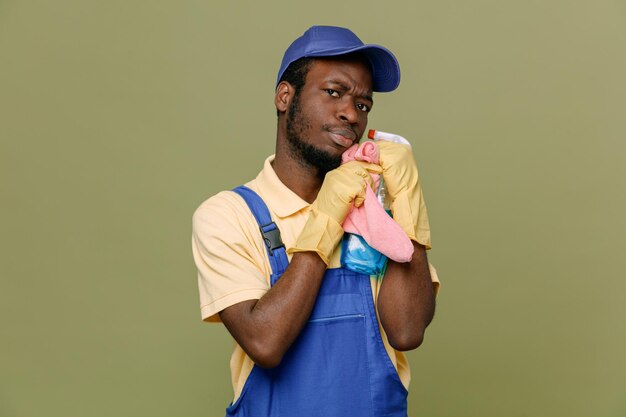 Greedy holding cleaning agent with rag young africanamerican cleaner male in uniform with gloves isolated on green background