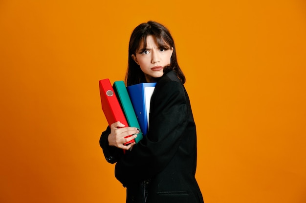 greedy holding folders young beautiful female wearing black jacket isolated on orange background