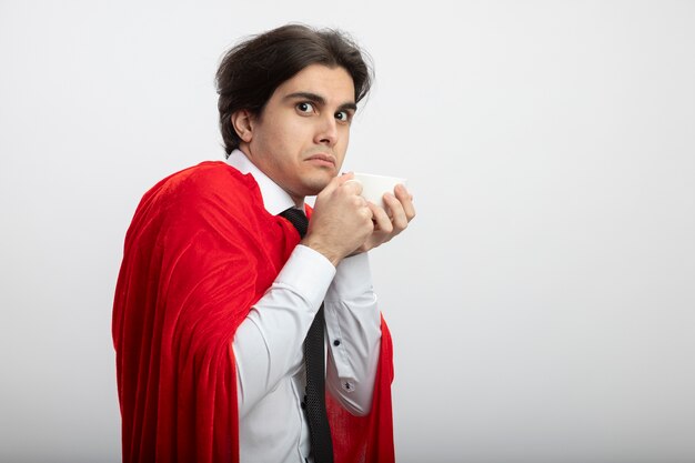 Greedy young superhero guy looking at camera wearing tie holding cup of tea isolated on white