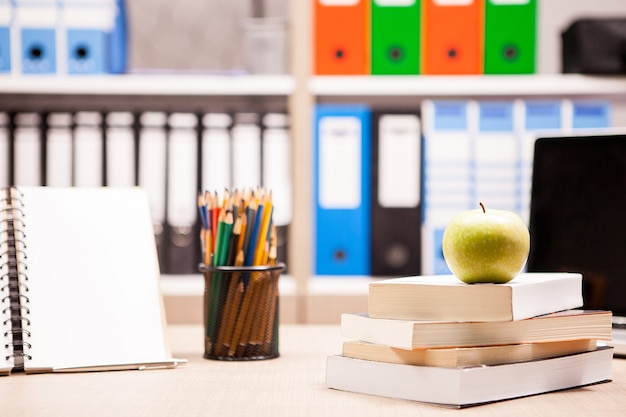 Free photo green apple on pile of books next to a notebook and pencils on table with a blurred white board in the back. school concept