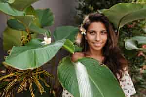 Free photo green-eyed brunette woman with flower in her hair looks into front, posing among large leaves of tropical plant