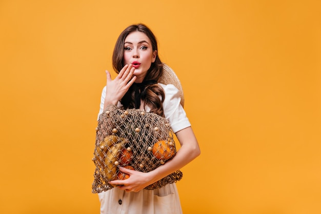 Free photo green-eyed lady in white cotton outfit with black bow is looking surprised at camera with string bag in her hands.