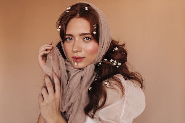 Green-eyed woman in light outfit coquettely looks at camera on beige background, playing with scarf.