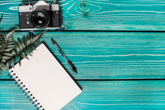 Free photo green fern leaves; spiral notepad; pen and camera against wooden turquoise background