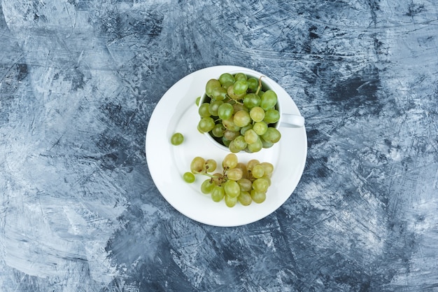 Green grapes in white cup and plate top view on a grungy plaster background