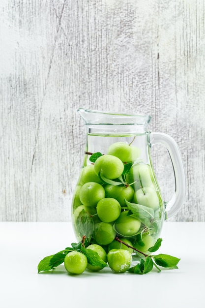 Free photo green plums with leaves in a glass jug on white and grungy wall, side view.