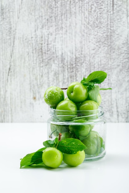 Free photo green plums with leaves in a glass mini jar on white and grungy wall, side view.