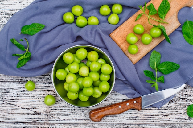 Free photo greengages with leaves in a metal saucepan and wood cutting board with knife flat lay on grey wood and picnic cloth