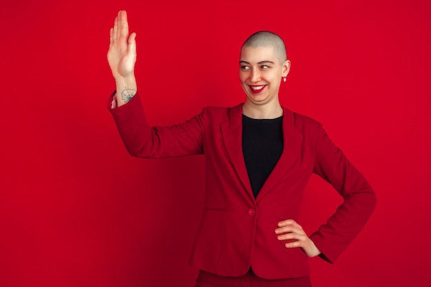 Greeting somebody. Portrait of young caucasian bald woman isolated on red wall. Beautiful female model in jacket. Human emotions, facial expression, sales, ad concept. Freaky.