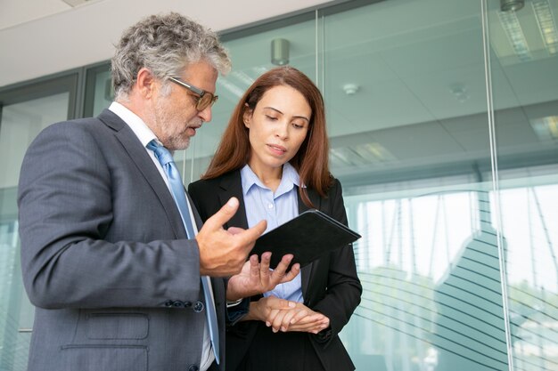 Grey-haired boss discussing with assistant, holding tablet and standing in conference room