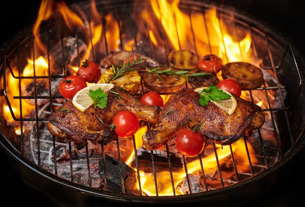 Free Photo grilled chicken legs on the flaming grill with grilled vegetables with tomatoes, potatoes, pepper seeds, salt.