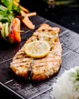 Free photo grilled fish fillet with lemon and vegetable sticks on a black steak board.