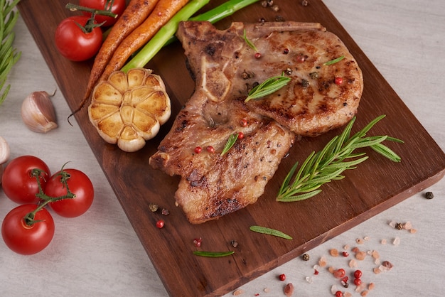 Grilled pork steak from a summer BBQ served with Vegetables, asparagus, baby carrots, fresh tomatoes and  spices. Grilled steak on wooden cutting board  on stone surface . top view.