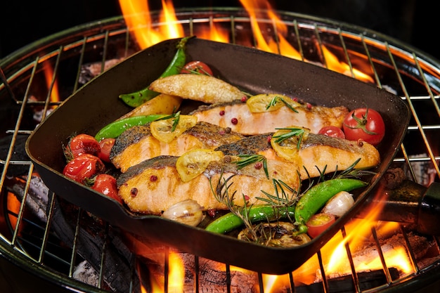 Grilled salmon fish with various vegetables on pan on the flaming grill pepper lemon and salt, herb decoration.