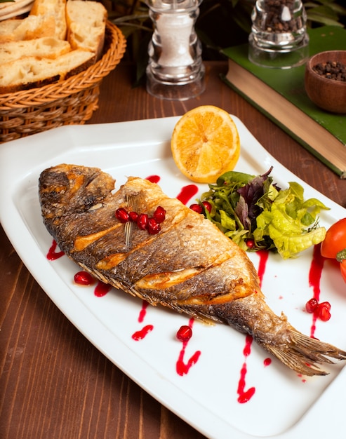 Grilled whole fish with yellow sauce, vegetable salad, lemon and pomegranate seeds in white plate