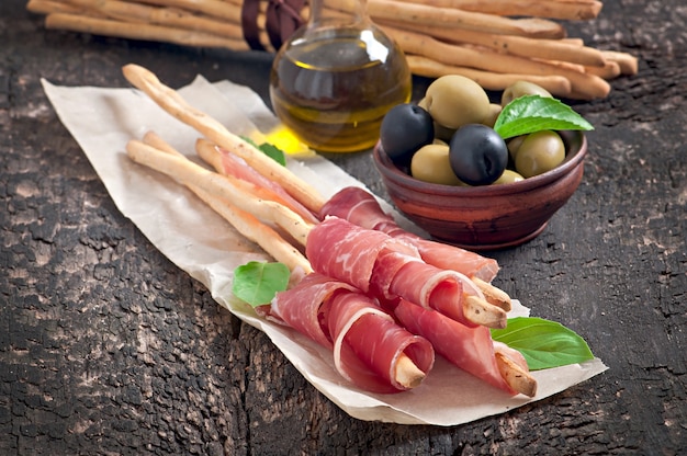 Grissini bread sticks with ham, olives, basil on old wood