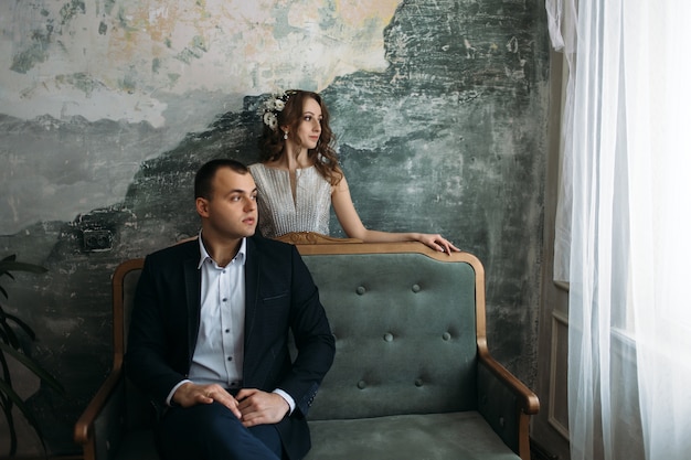 Free photo the groom sits on the sofa and bride stands near her husband