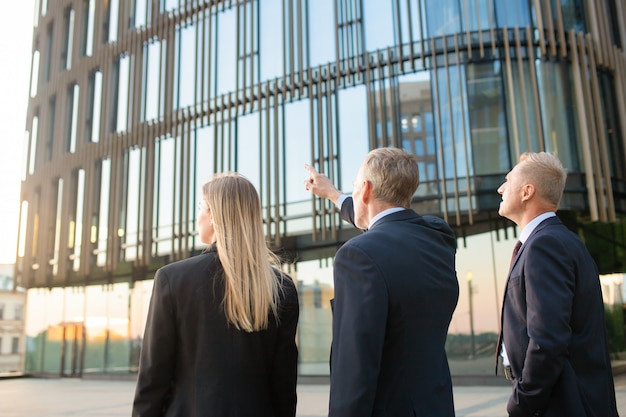 Free photo group of business partners in formal suits pointing at office building, meeting outdoors, discussing real property. back view. commercial real estate concept