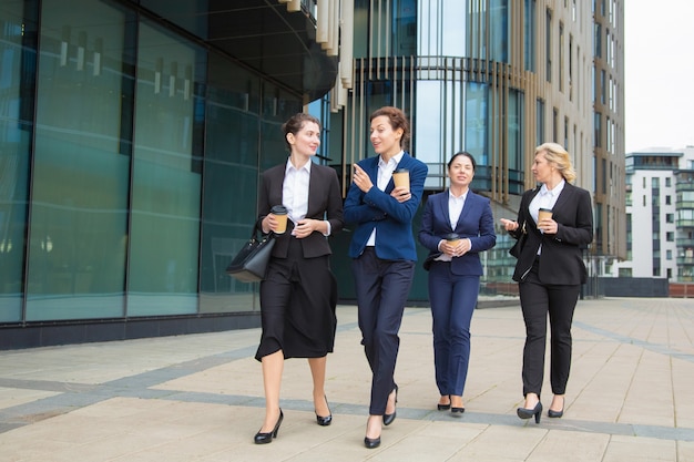 Free Photo group of female coworkers walking with takeaway coffee outdoors, talking, smiling. full length, front view. work break concept