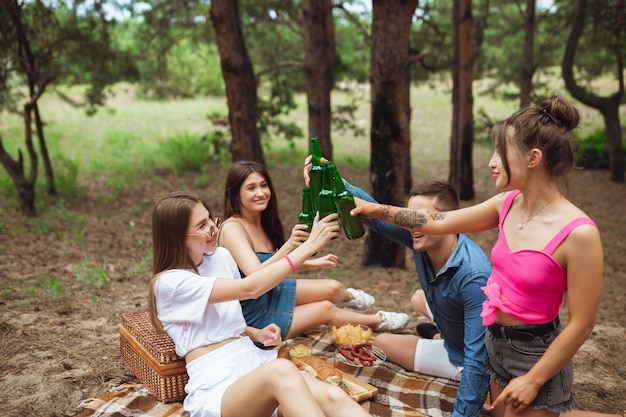 Group of friends clinking beer bottles during picnic in summer forest