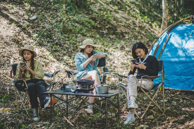 Group of girl friends sitting on camping chair and use their smartphone ignoring each other while camping in park