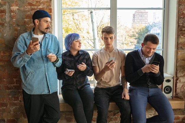 Group of happy caucasian young people standing behind the window. Sharing a news, photos or videos from smartphones, talking or playing games and having fun. Social media, modern technologies.