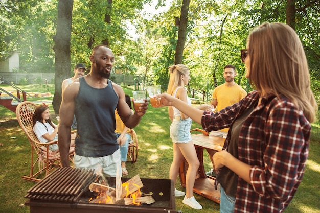 Group of happy friends having beer and barbecue party at sunny day. Resting together outdoor in a forest glade or backyard. Celebrating and relaxing, laughting. Summer lifestyle, friendship concept.