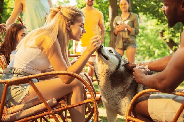 Group of happy friends having beer and barbecue party at sunny day. Resting together outdoor in a forest glade or backyard. Celebrating and relaxing, laughting. Summer lifestyle, friendship concept.