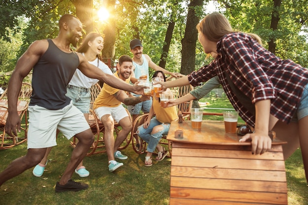 Group of happy friends having beer and barbecue party at sunny day. Resting together outdoor in a forest glade or backyard