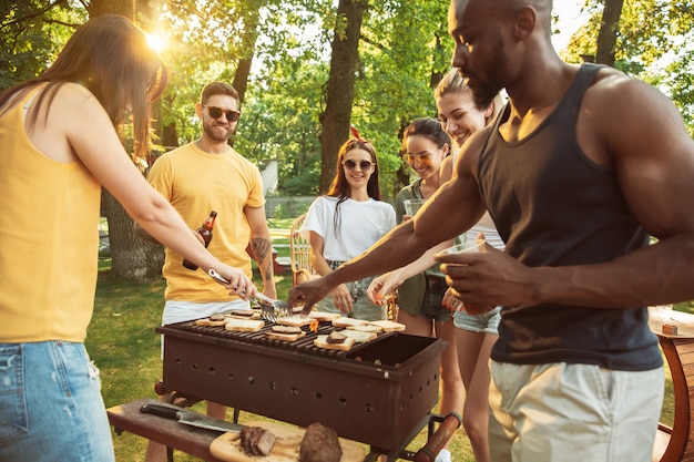 Group of happy friends having beer and barbecue party at sunny day. Resting together outdoor in a forest glade or backyard