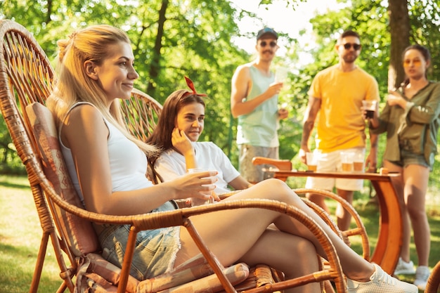 Group of happy friends having beer and barbecue party at sunny day. Resting together outdoor in a forest glade or backyard