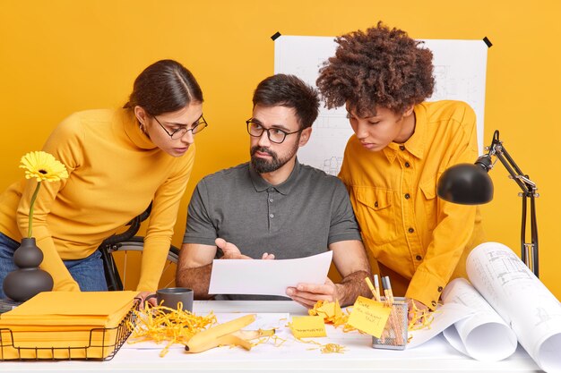 Group of international architects discuss ideas for engineering project enjoy working process together pose at desktop concentrated at paper yellow wall. Diverse colleagues inn office.