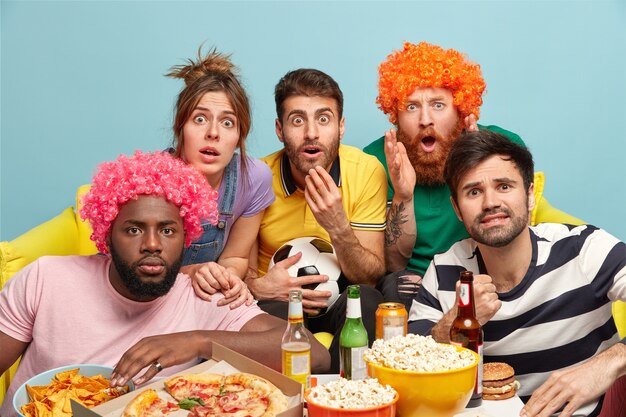 Group of multiethnic friends stare, have bated breath while watch very exciting football game, sit sofa near table with pizza, beer and popcorn isolated on blue wall. Crazy emotional reaction