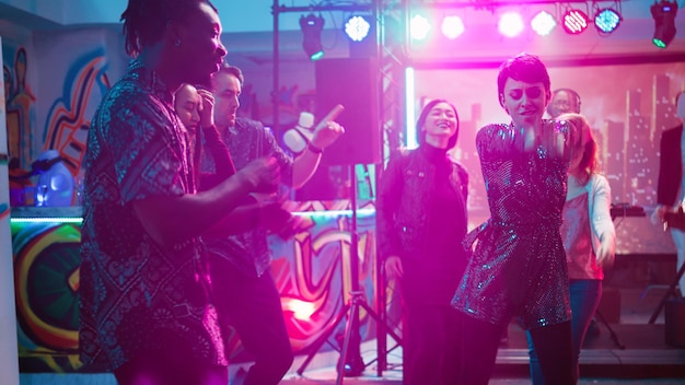 Free Photo group of people enjoying disco party, dancing on club music at social gathering. young men and women showing dance moves on dance floor, partying with stage lights. handheld shot.