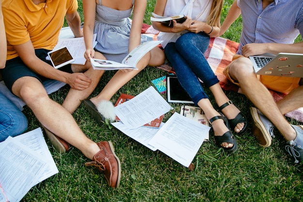 Free photo group of students with books and tablet are studying outdoors together, sitting on grass.