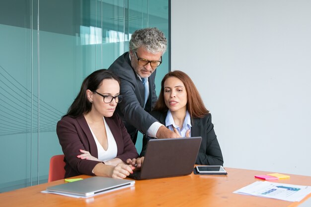 Group of top managers watching and discussing project presentation on laptop, male executive pointing at display, while female managers explaining details