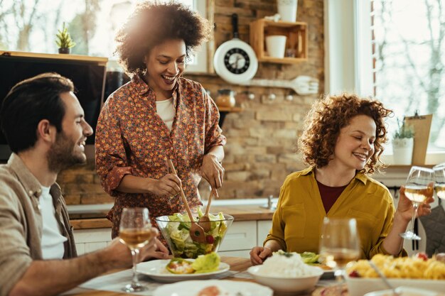 Group of young happy friends enjoying in lunch Focus is on African American woman serving salad at dining table