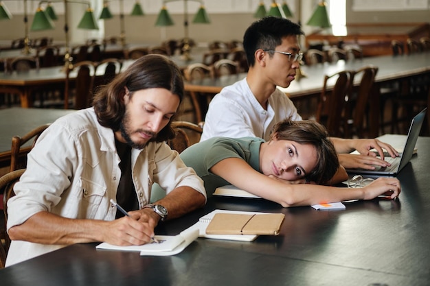 Group of young multinational students studying together in library of university Attractive tired student girl lying on table while thoughtfully looking in camera