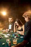 Free photo group of young rich people is playing poker in the casino. two men in business suits and a young woman in a black dress. smoke. casino. poker