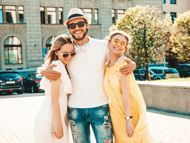 Group of young three stylish friends posing in the street. Fashion man and two cute girls dressed in casual summer clothes. Smiling models having fun in sunglasses.Cheerful women and guy going crazy