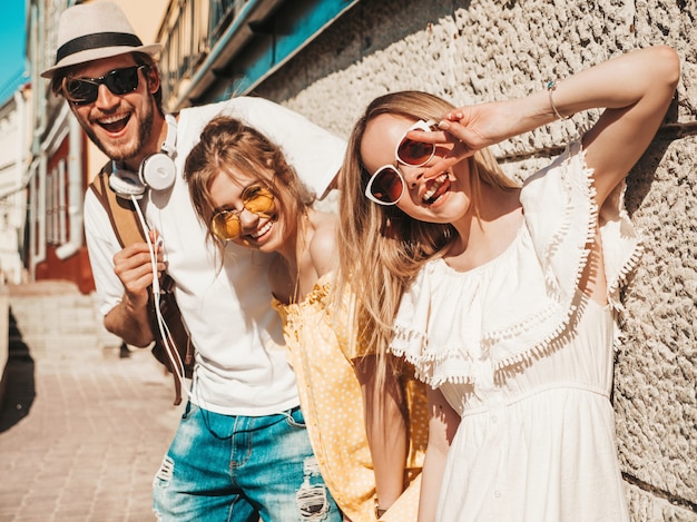 Group of young three stylish friends posing in the street. Fashion man and two cute girls dressed in casual summer clothes. Smiling models having fun in sunglasses.Cheerful women and guy outdoors