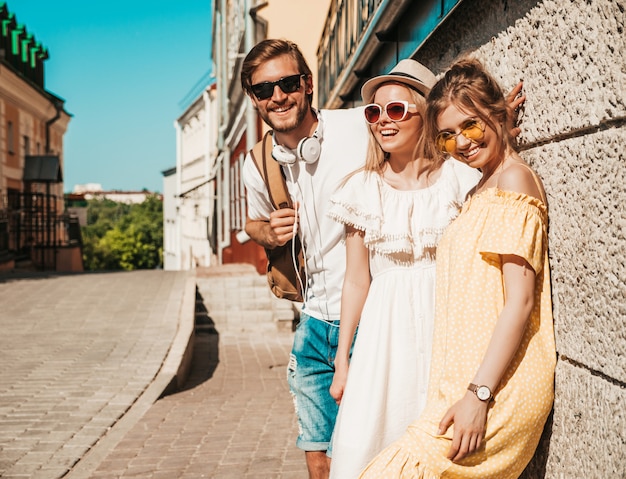 Group of young three stylish friends posing in the street. Fashion man and two cute girls dressed in casual summer clothes. Smiling models having fun in sunglasses.Cheerful women and guy outdoors