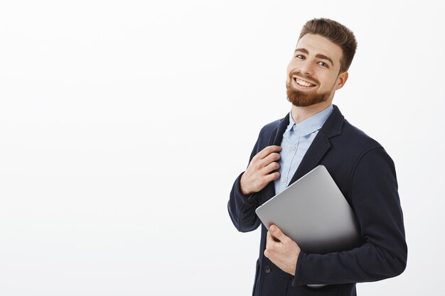 Guy can deal with any task feeling self-assured and pleased touching suit holding laptop in arm standing half-turned over gray wall gazing delighted and satisfied with own successful plan