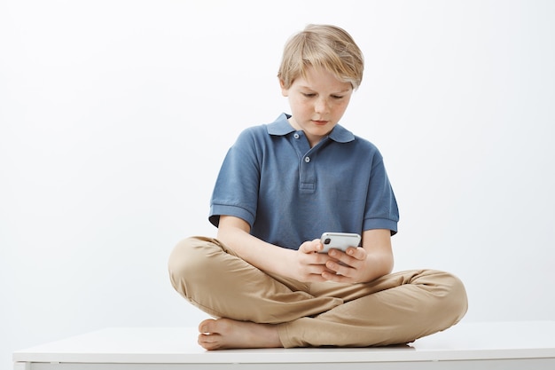 Guy feeling intense playing favorite game in smartphone. Serious pretty little boy with blond hair sitting on floor with feet crossed, holding telephone and gazing at device screen