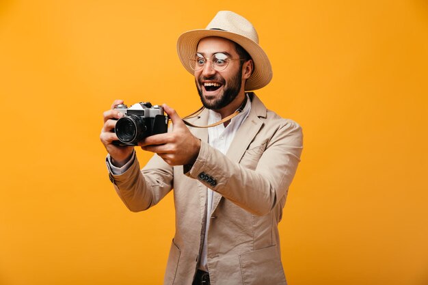 Guy in hat and beige jacket takes photos on retro camera on orange background