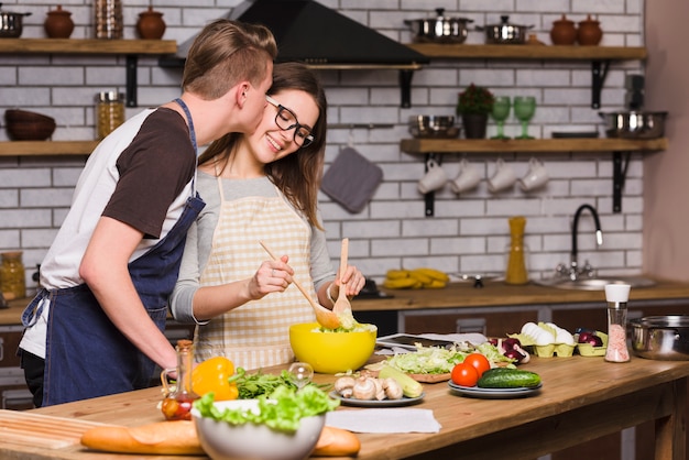 Free photo guy kissing young woman mixing salad in kitchen